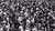 A black and white image featuring hundreds of people standing in a large crowd